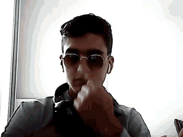 a young man wearing sunglasses and ear buds holds his hand to his mouth