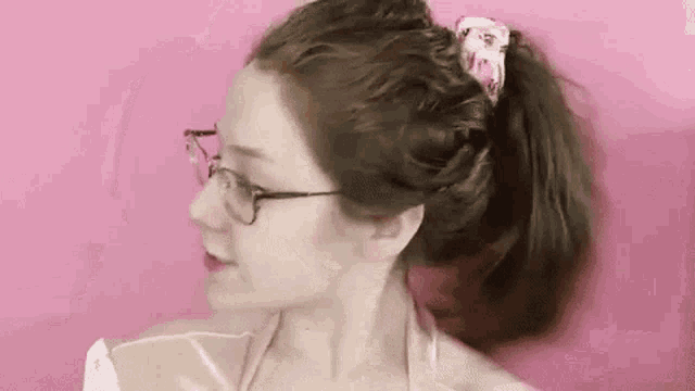 a woman wearing glasses and a pink scrunchie is holding her hair in a ponytail .