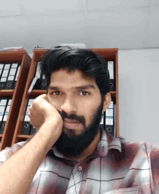 a man with a beard is sitting in front of a shelf with binders on it