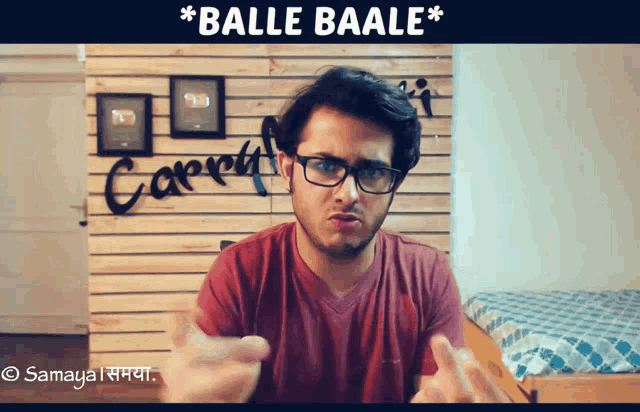 a man wearing glasses and a red shirt stands in front of a wall that says " balle baale "