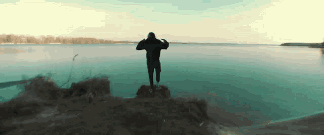 a man in a black hoodie stands on a rock overlooking a body of water