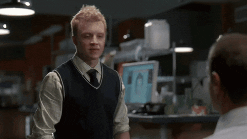 a man in a sweater vest and tie stands in front of a computer monitor