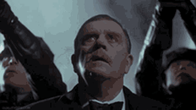 a man in a tuxedo and bow tie is standing in front of a group of men holding up their arms .