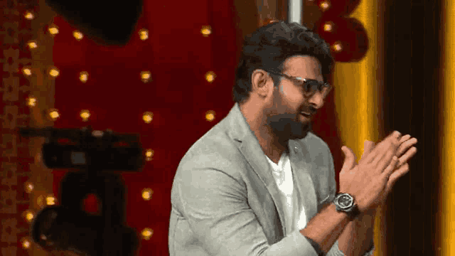 a man in a suit and glasses is clapping his hands in front of a camera .