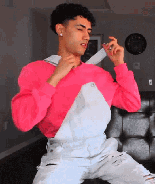a young man wearing a pink sweater and white overalls is sitting on a couch