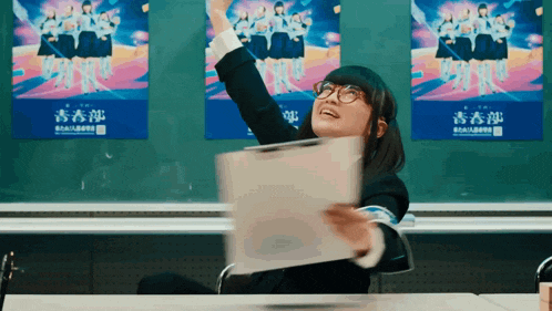 a girl sitting at a desk with her arms in the air in front of a blackboard with chinese writing