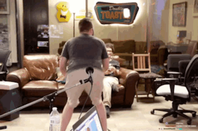 a man is standing in front of a microphone in a living room while another man sits in a chair .