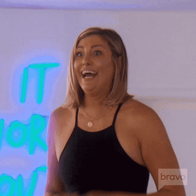 a woman in a black tank top is smiling in front of a neon sign that says " it 's more than "