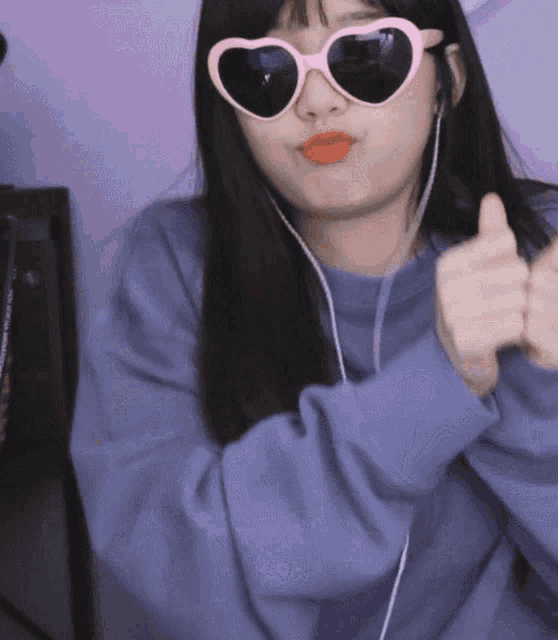 a woman wearing heart shaped sunglasses and earbuds gives a thumbs up