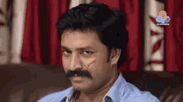 a man with a mustache is sitting on a couch in front of a curtain that says flowers