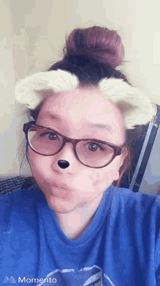 a woman wearing glasses and a blue shirt is taking a selfie with a momento logo in the corner