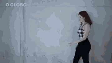 a woman in a plaid top and black pants is walking against a gray wall .