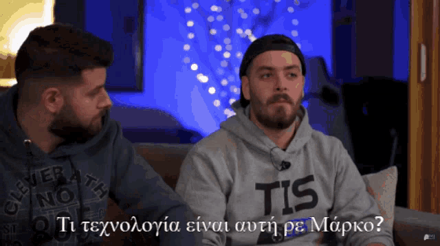 two men are sitting on a couch and one has a sweatshirt that says tis