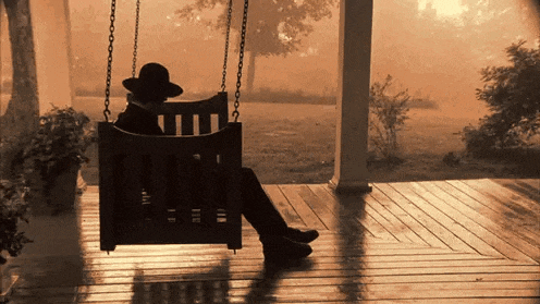 a man in a hat sits on a porch swing in the evening