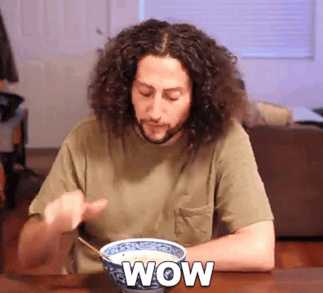a man with curly hair is sitting at a table with a bowl of food and the word wow is on the table