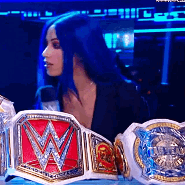 a woman with blue hair is standing next to a bunch of wrestling championship belts .