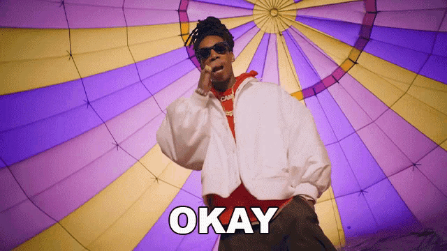 a man in a white jacket stands in front of a purple and yellow hot air balloon and the word okay is on the screen