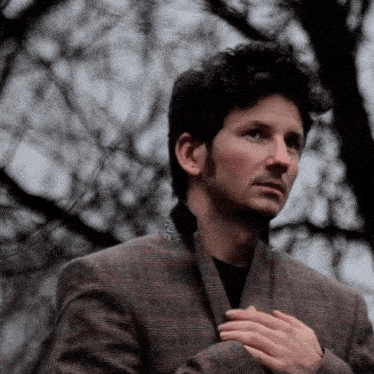 a man in a plaid jacket has his hands folded in front of his chest
