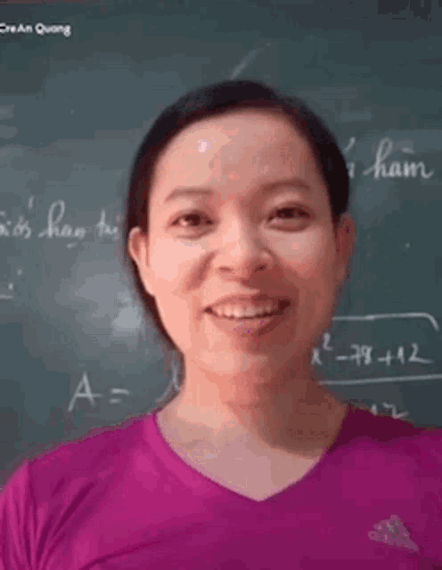 a woman in a pink shirt is smiling in front of a blackboard with writing in a foreign language