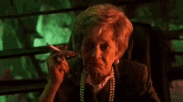 an elderly woman is smoking a cigarette in a dark room and giving the middle finger .
