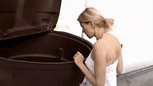 a woman in a white tank top is standing next to a black trash can with the lid open