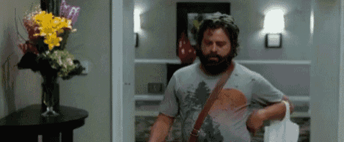 a man with a beard is standing in front of a vase of flowers in a hallway .