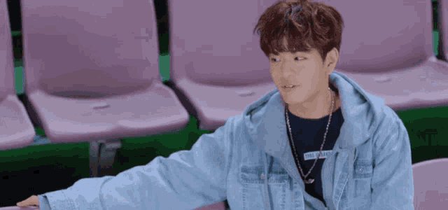 a young man wearing a denim jacket and a necklace is sitting on a bench