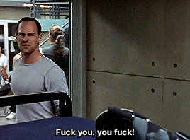 a man in a grey shirt is standing in a room and says " fuck you you fuck "