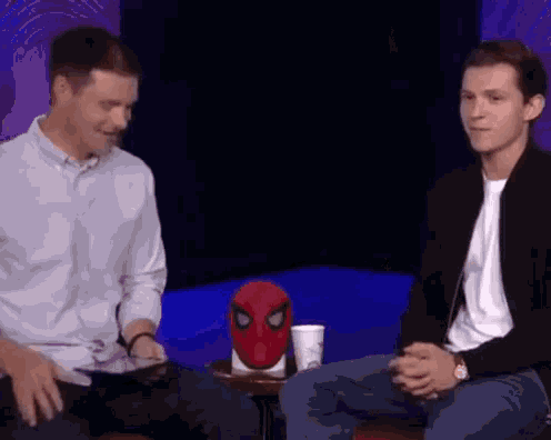 two men are sitting next to each other on a table with a spider-man mask in the background .