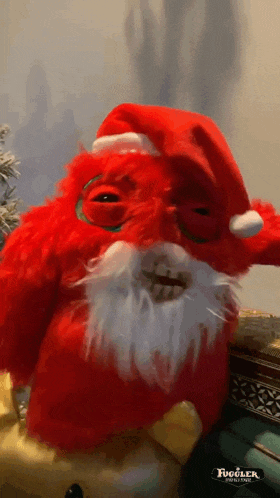 a red stuffed animal with a santa hat and beard is sitting on a table with a fuddler logo on the bottom right