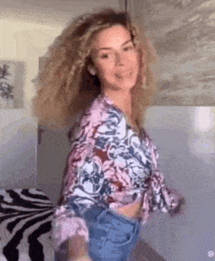 a woman with curly hair is standing in a room wearing a floral shirt and jeans .