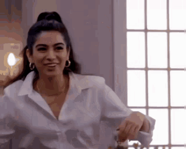a woman in a white shirt and hoop earrings is standing in front of a window and smiling .