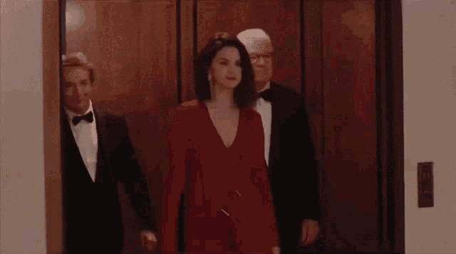 a woman in a red dress is standing next to two men in tuxedos in an elevator .