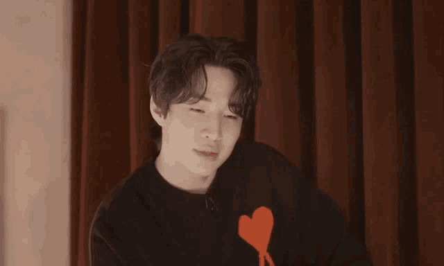 a young man wearing a black sweater with a heart on it is making a heart shape with his finger .
