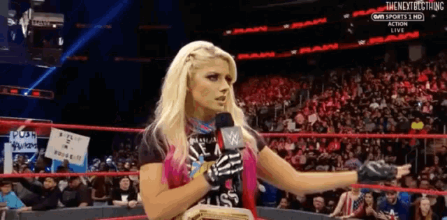 a woman holding a microphone in a wrestling ring with the next best thing written on the bottom right