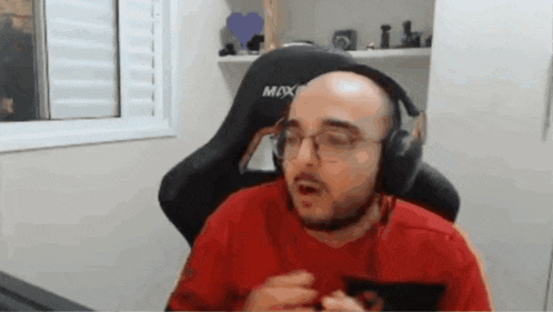 a bald man wearing headphones and a red shirt is sitting in a gaming chair .