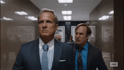 two men in suits and ties are walking down a hallway in an office .