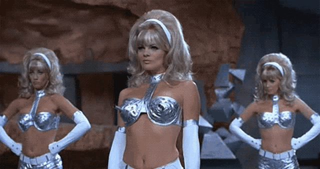 a woman in a silver bra and white gloves is standing next to two other women in silver costumes .