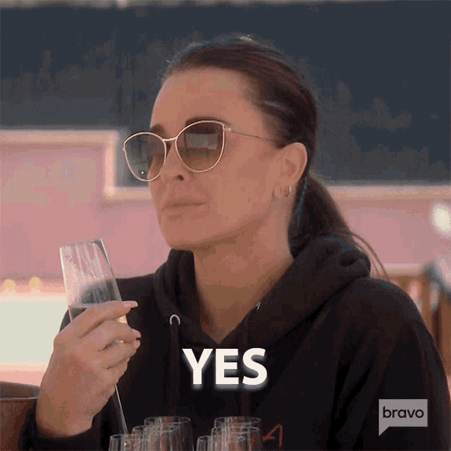 a woman wearing sunglasses is holding a glass of wine and says " yes "