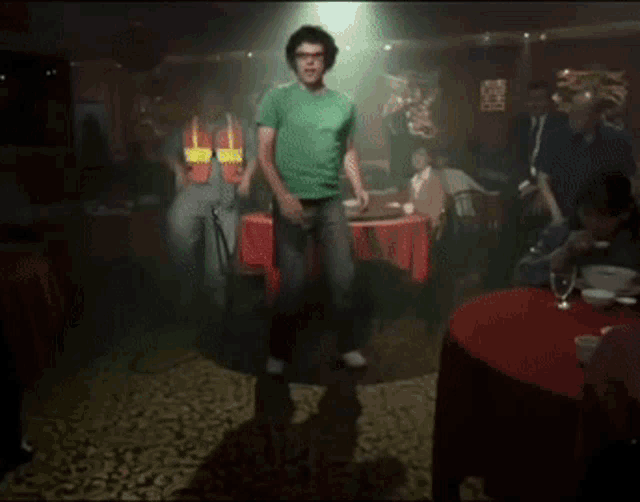 a man in a green shirt is dancing in a room with people sitting at tables
