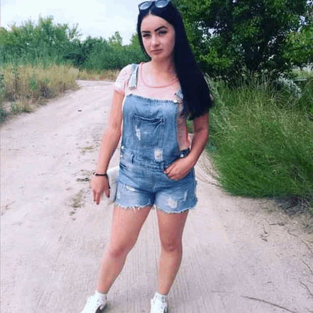 a woman is standing on a dirt road wearing overalls .
