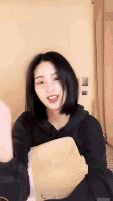 a woman in a black hoodie is holding a pillow and making a funny face