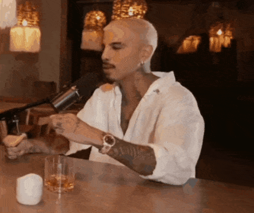 a man is sitting at a table talking into a microphone while holding a glass of whiskey .