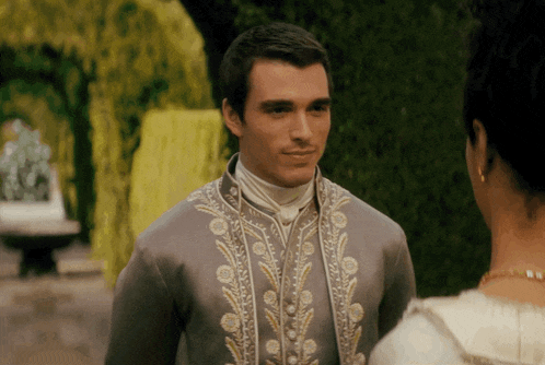 a man in a grey jacket with gold embroidery looks at a woman