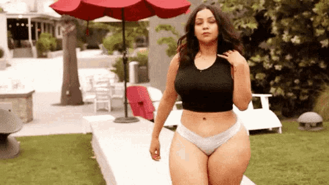a woman in a black crop top and white underwear is standing in front of a red umbrella in a backyard .