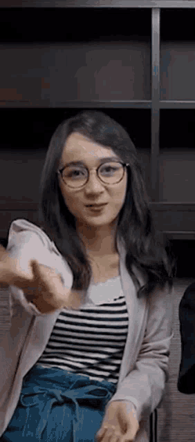 a woman wearing glasses and a striped shirt is sitting in a chair and giving a thumbs up .