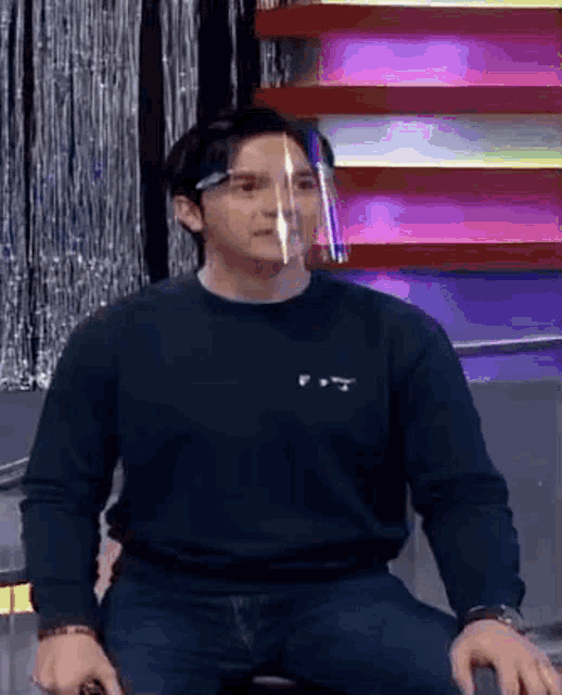 a man wearing a face shield and a black sweater is sitting in a chair .