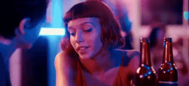 a woman is sitting at a bar with two bottles of beer and talking to a man .