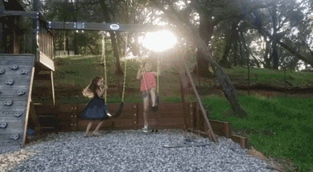 two girls are playing on a swing set with the sun shining through the trees in the background