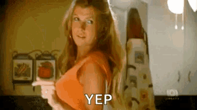 a woman in an orange top is standing in a kitchen and pointing at her breast .